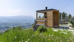 Een houten hutje met een glazen raam, gelegen op een grasheuvel met uitzicht op een bergachtig landschap.