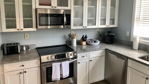 Una cocina con armarios blancos, electrodomésticos de acero inoxidable, encimera de granito y una ventana con persianas.