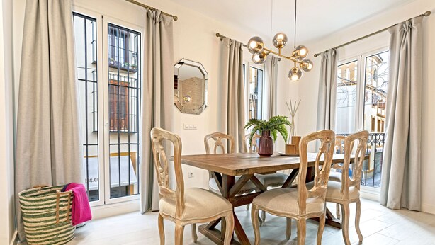 A dining area with a wooden table, six chairs, a chandelier, and large windows with curtains. - Genteel Home Feria II (Sevilla)