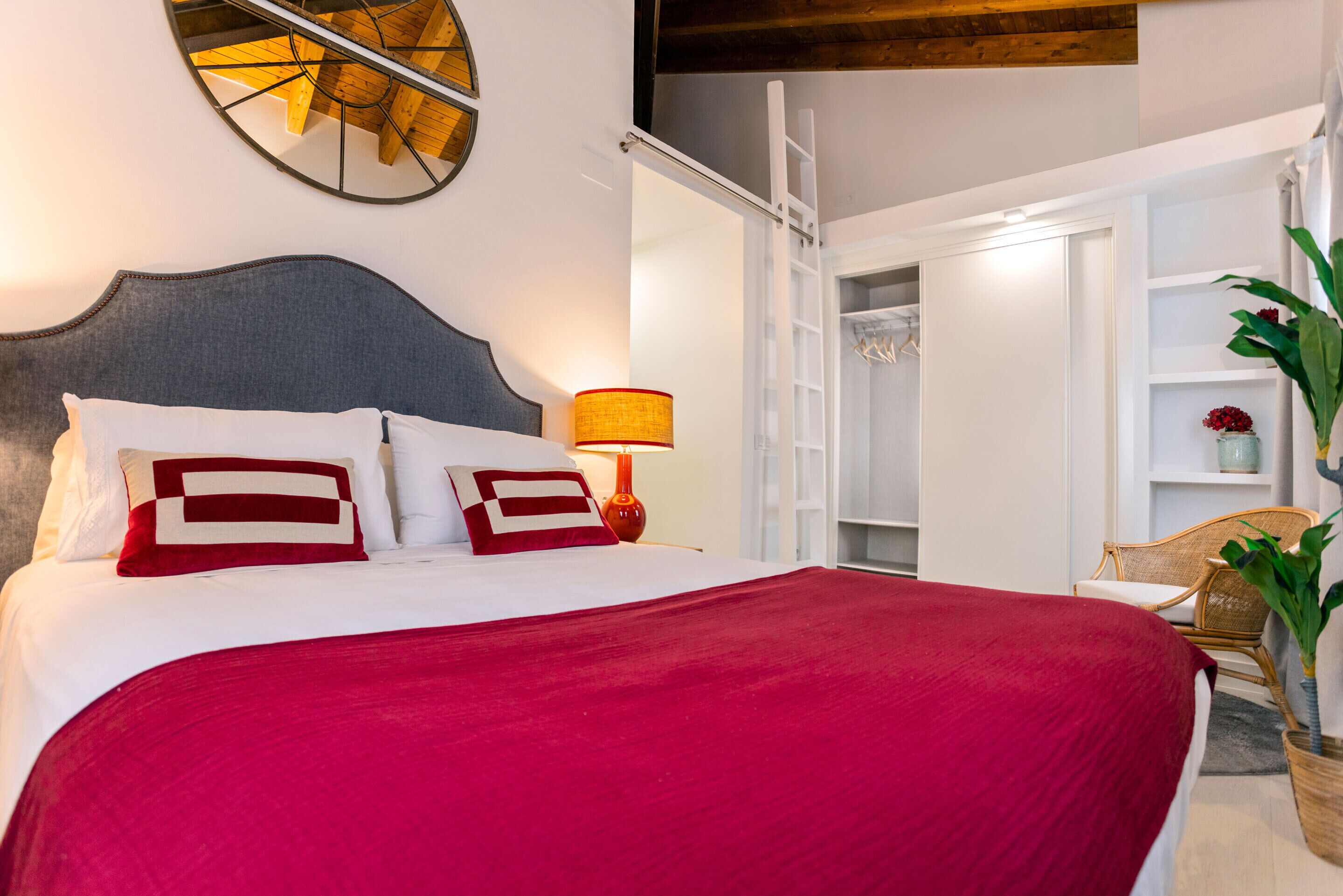 Een opgemaakt bed met een rode deken en kussens, een bedlampje en een houten hoofdbord.