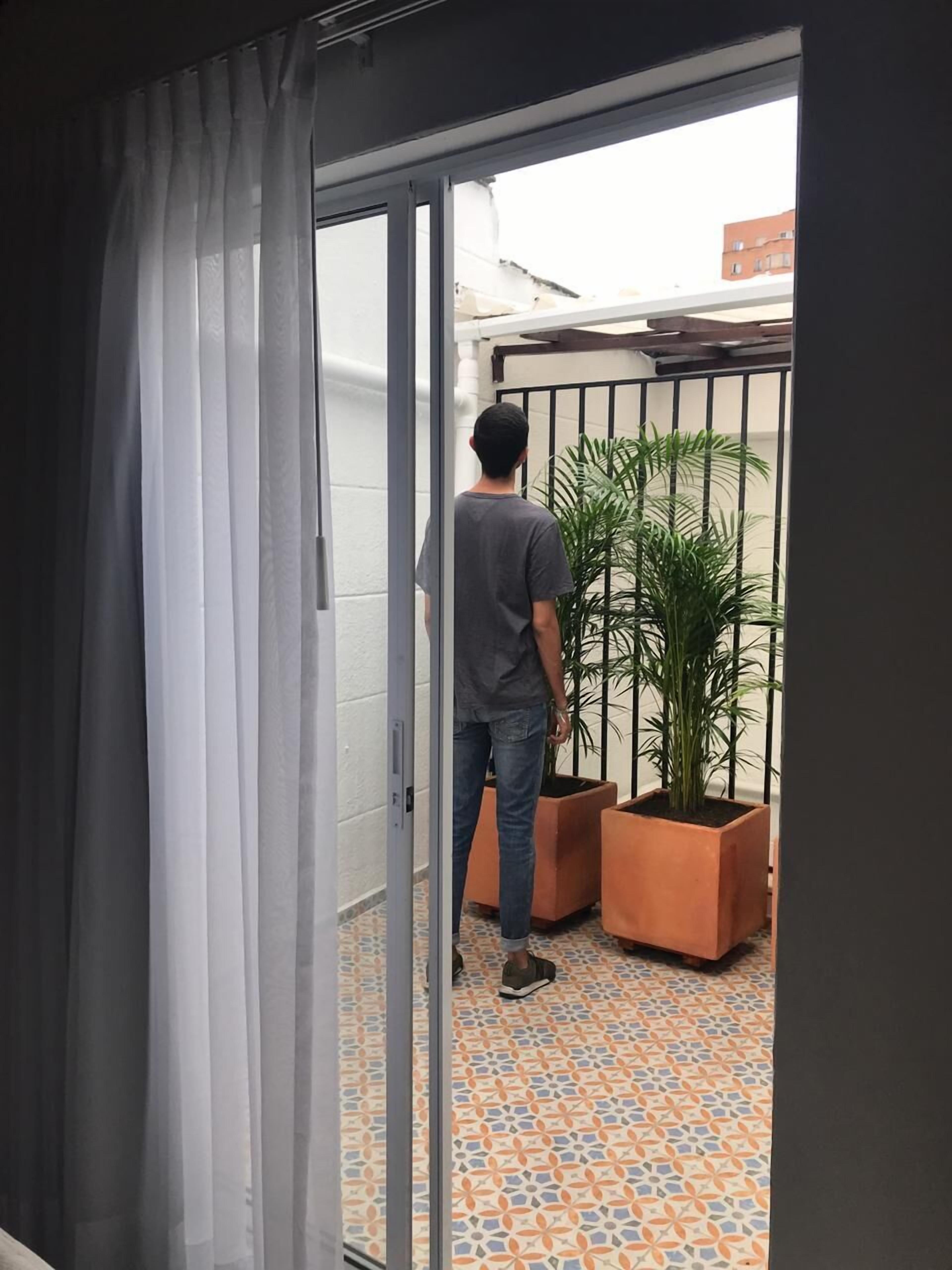 Un homme se tient dans l’encadrement d’une porte, devant une porte-fenêtre coulissante, et regarde vers l’extérieur. On aperçoit deux plantes en pot et un portail en métal dans l’espace extérieur.