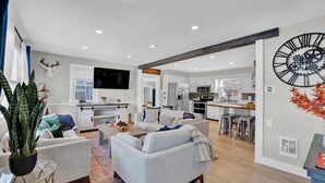 A modern living room with a sofa, a coffee table, and a TV. There is a kitchen area with a fridge, microwave, and a clock on the wall.