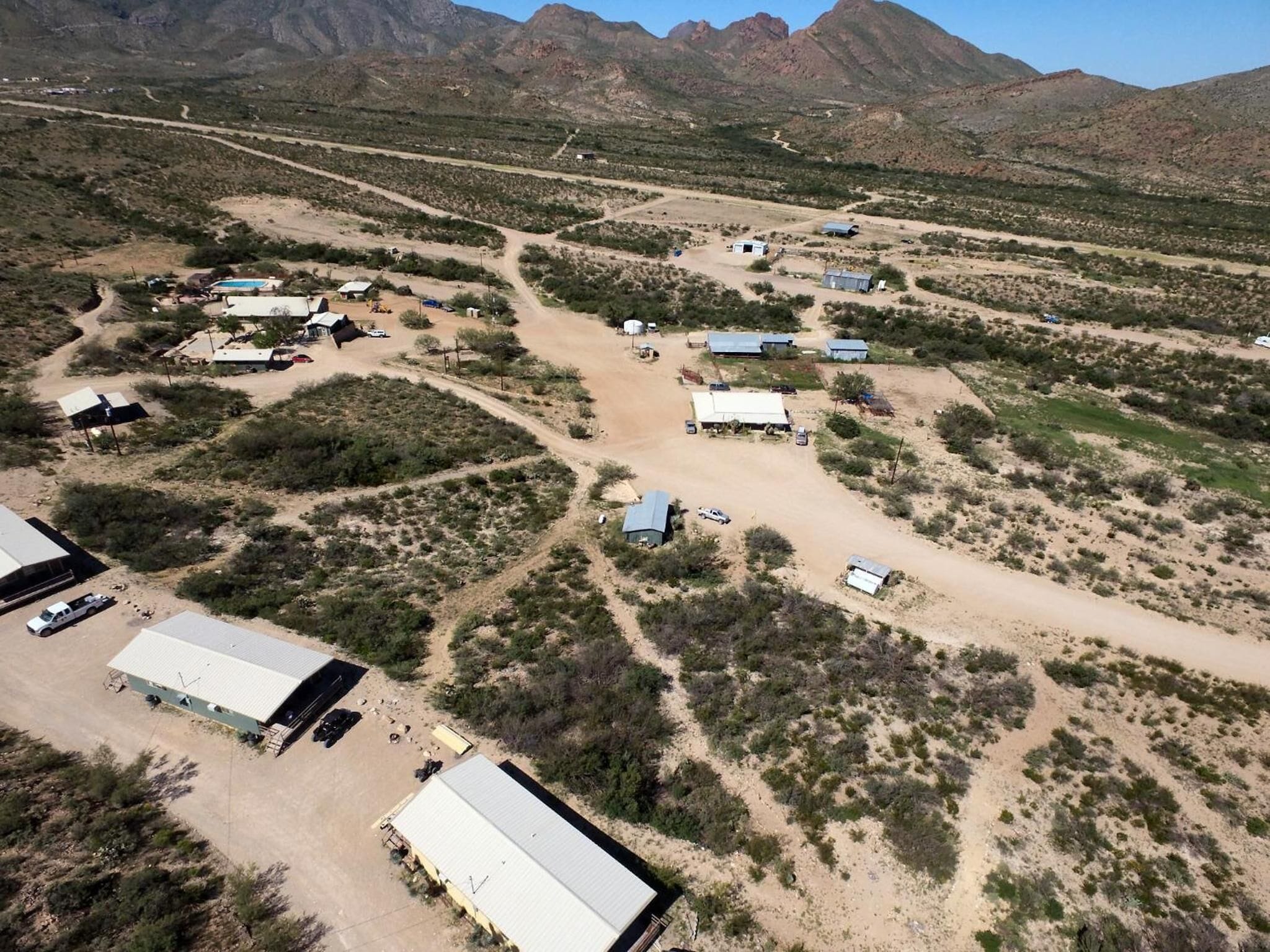 An aerial view of a rural area with several buildings, including a large warehouse and smaller structures, surrounded by sparse vegetation and mountains in the background.