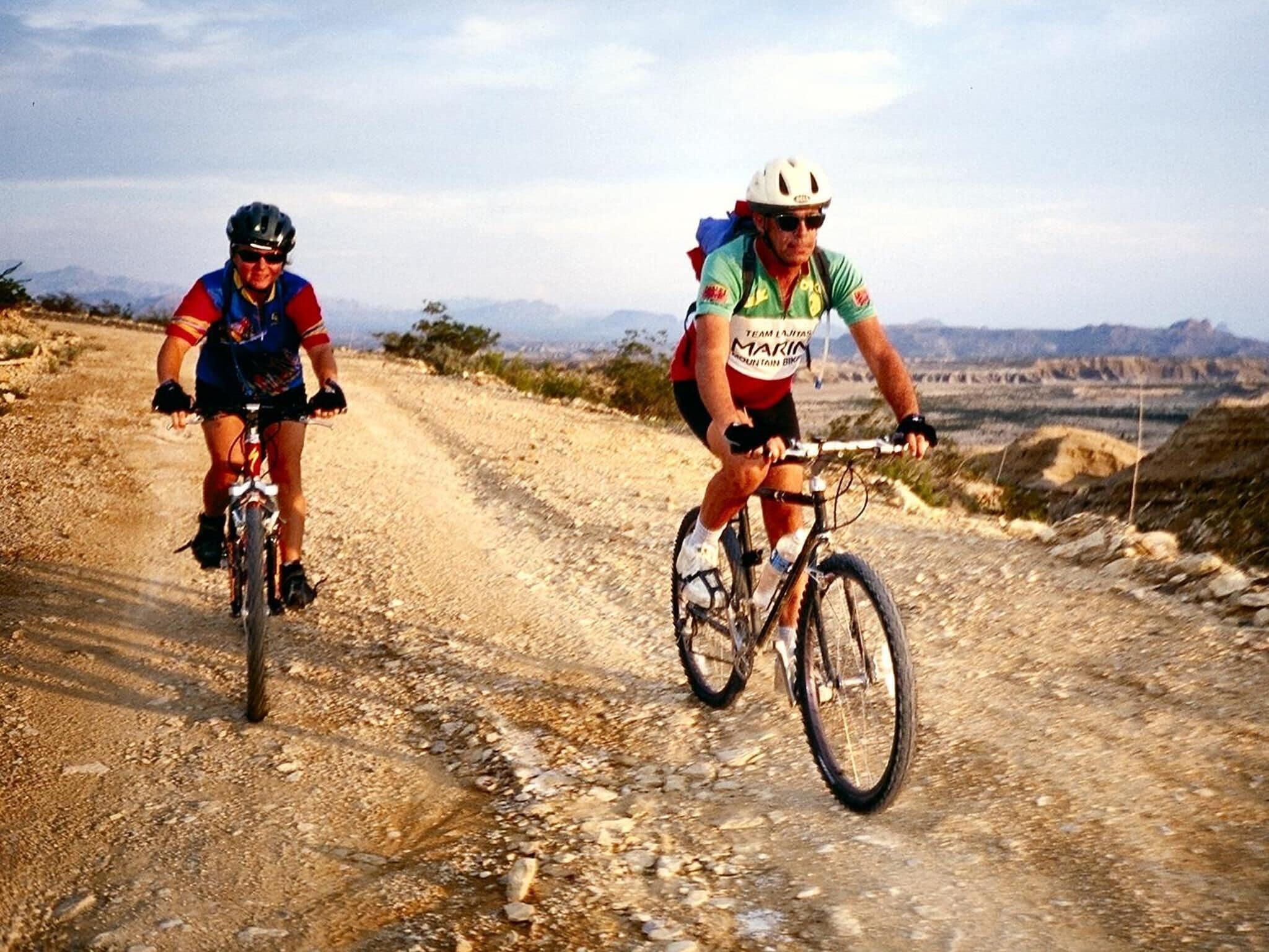 Deux cyclistes sur un chemin de terre, avec un paysage montagneux en arrière-plan.