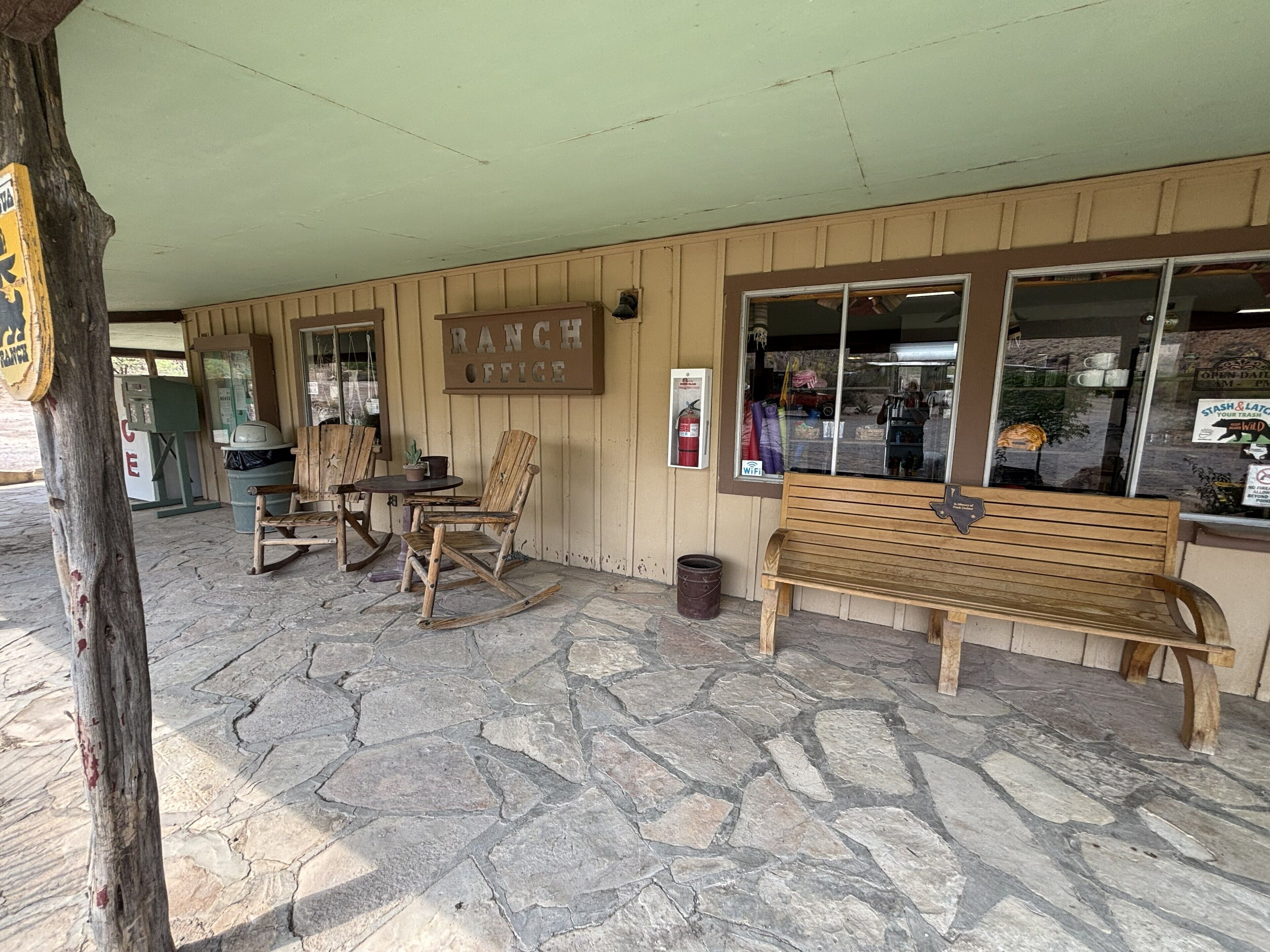Un espace extérieur rustique aménagé avec des bancs en bois et une table, une enseigne indiquant « BUREAU DU RANCH » et un sol pavé de pierres.