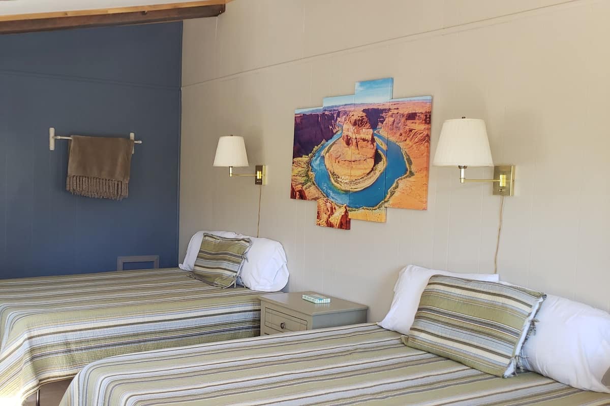 A room with two beds, a sink, and a mirror. There are wall-mounted lamps and a painting.