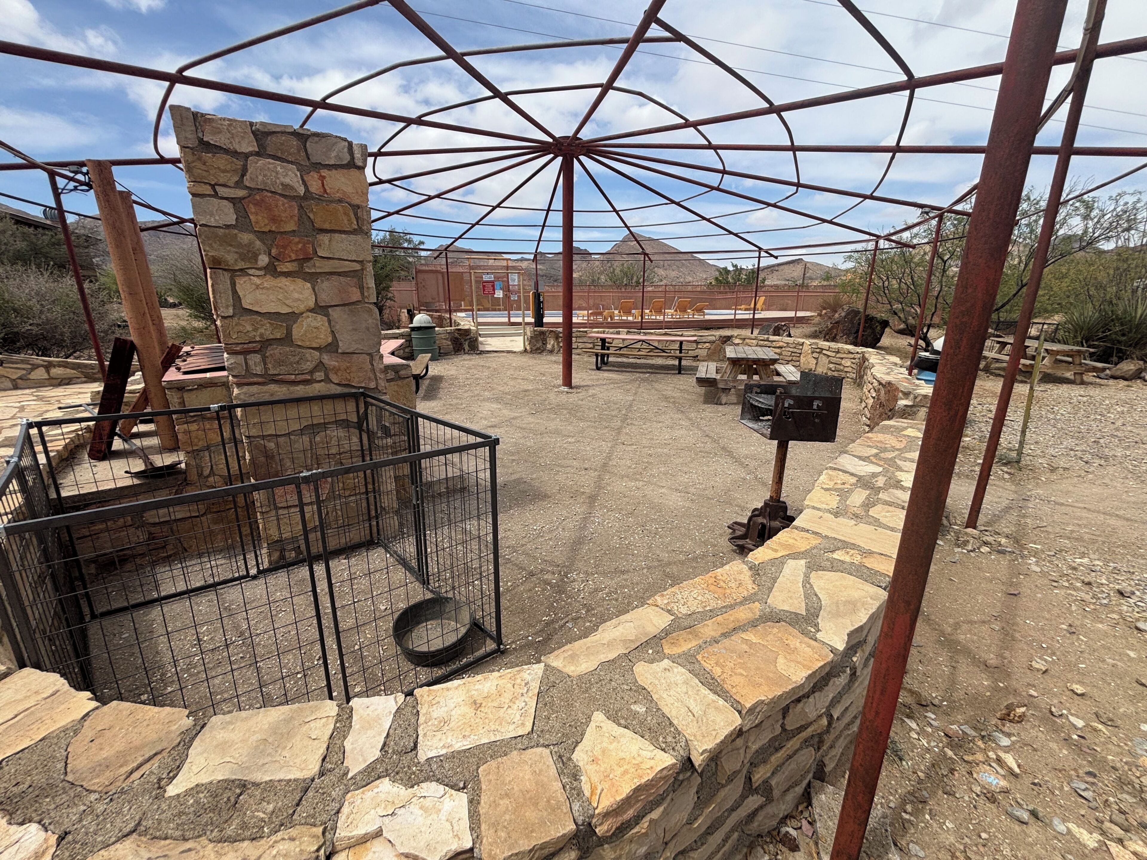 Un espacio al aire libre con paredes de piedra, una parrilla de metal, mesas de picnic y una chimenea de piedra.