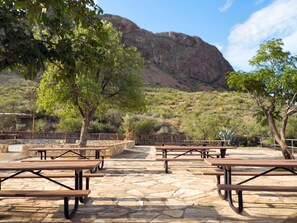An outdoor patio with stone flooring and wooden picnic tables, surrounded by trees and a mountainous backdrop.