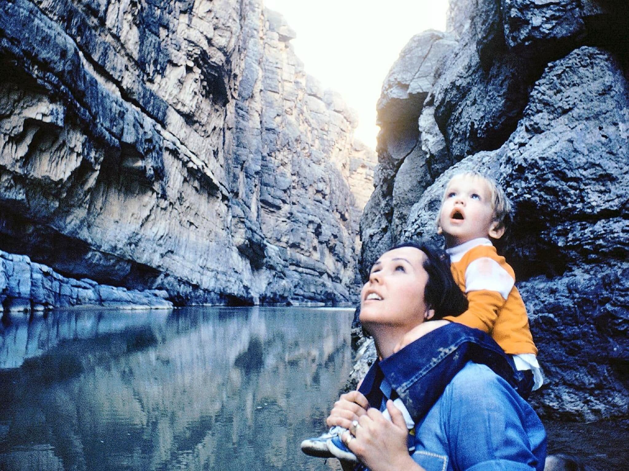 Eine Person hält ein Kind in einer felsigen Schlucht mit einem ruhigen Fluss.