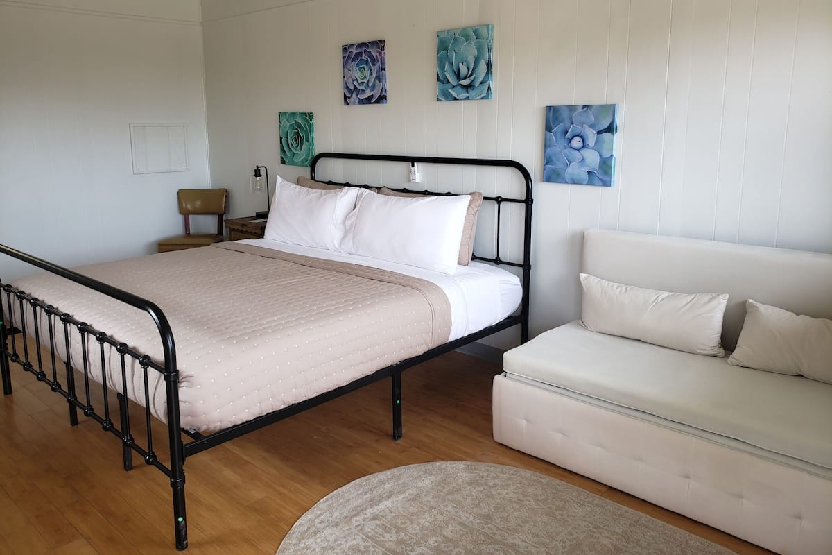 A bedroom with a bed, a nightstand, a lamp, a chair, a sofa, and three wall art pieces.
