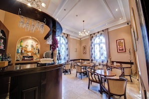 A bar area with a counter, shelves with bottles, and a chandelier. There are dining tables with chairs in the background.