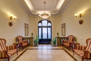 A lobby with a chandelier, two armchairs, a large arched doorway, and a tiled floor.