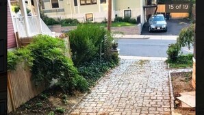 Een geplaveid pad in een woonwijk, met aan één kant een houten hek en groen, en aan de andere kant een geparkeerde auto.