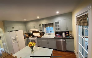 A kitchen with light blue cabinets, a white refrigerator, a stove, a sink, and a dishwasher. There is a window with a plaid curtain, and a vase with yellow flowers on the table. - Victorian house, clean comfortable and well located (Boston)