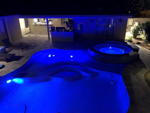A hot tub with blue lighting, a patio area with lounge chairs, and a building with a glass door in the background. - Scottsdale McCormick Ranch Oasis – Heated Pool, Slide, Spa – Walk to Restaurants (Scottsdale)