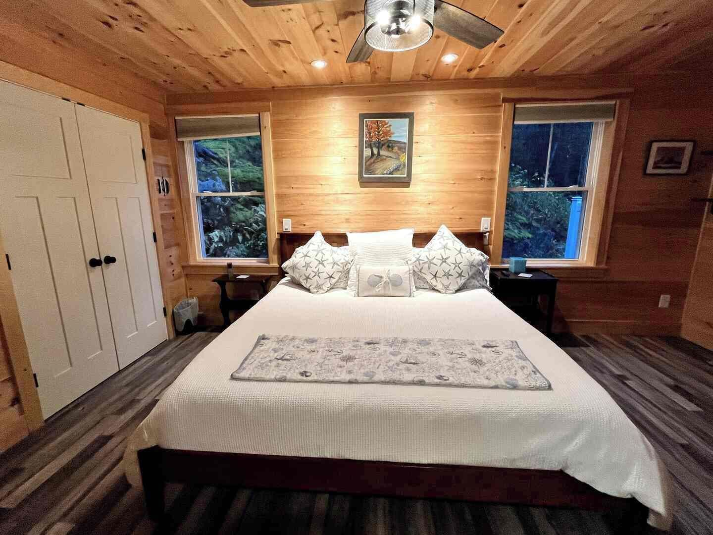 A bedroom with a large bed, two windows, a ceiling fan, and a painting on the wall.