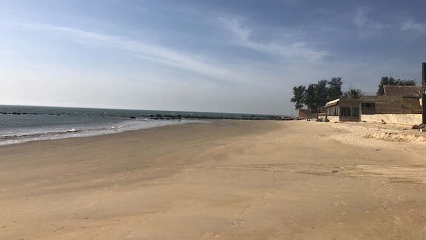 Ein weiter Sandstrand, bewölkter Himmel und ruhiges Meer. Im Hintergrund sind Gebäude und Bäume zu sehen.