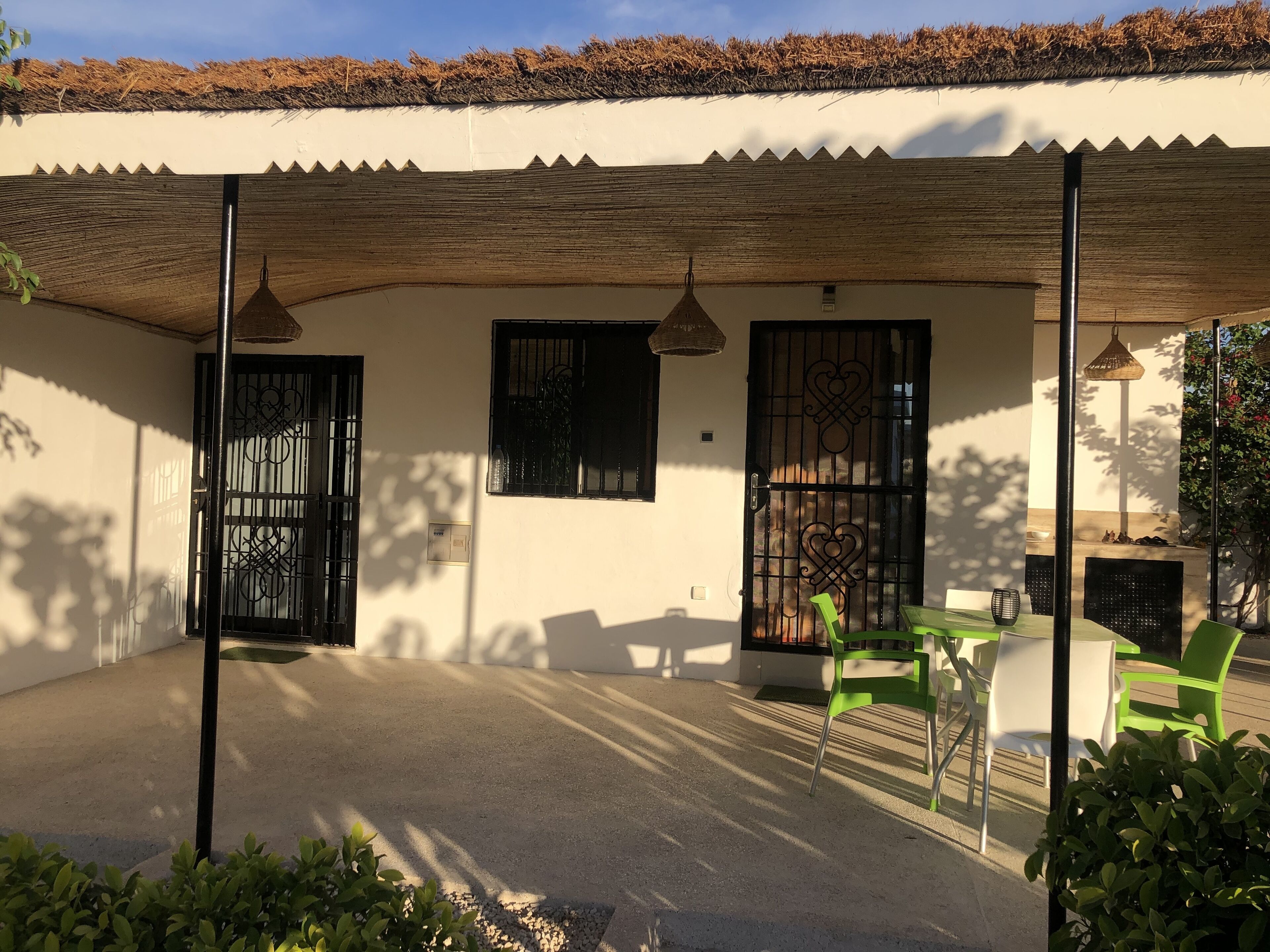 Un espace extérieur abrité, doté d’un sol en béton, de deux fenêtres avec des barreaux en métal et d’une porte. On y trouve des chaises vertes et blanches ainsi qu’une table.