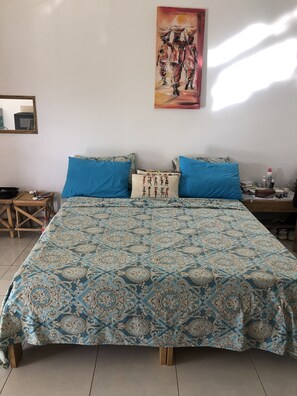 A bedroom with a patterned bedspread, two blue pillows, a decorative pillow with a figure design, a painting on the wall, a wooden nightstand, and a mirror. - La Maison Aziz, air-conditioned & quiet. A 15-minute walk from the center of Somone (Somone)