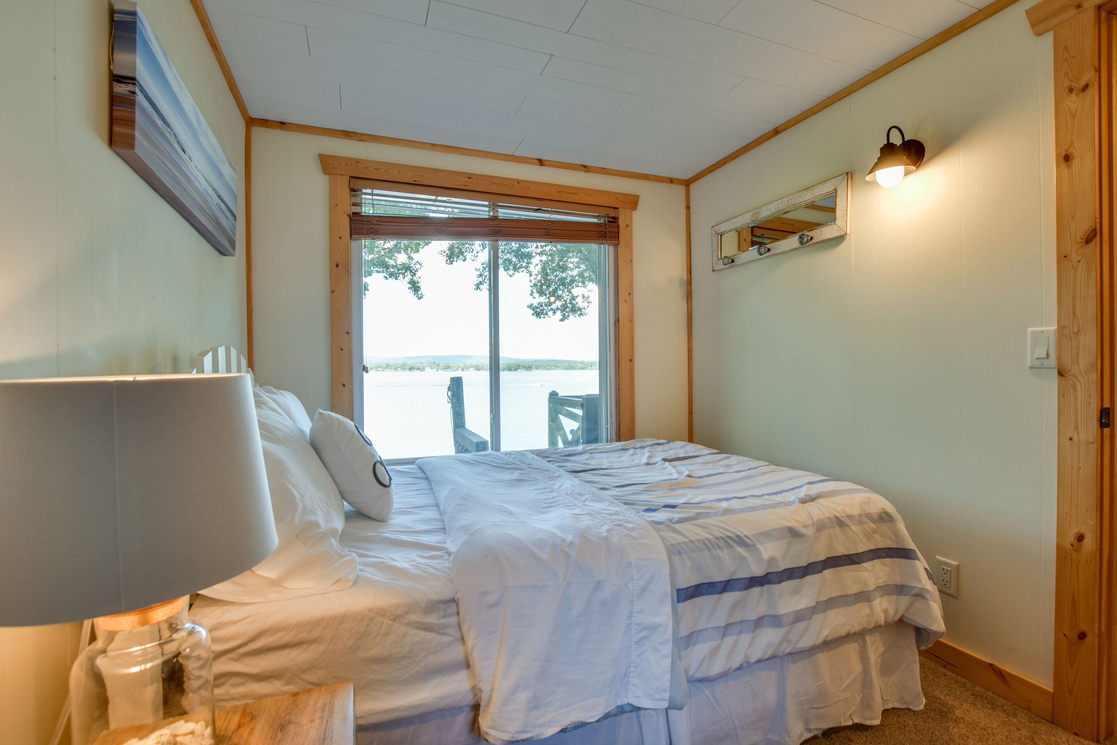 A bedroom with a bed, a lamp, a window with blinds, a mirror, and a door.