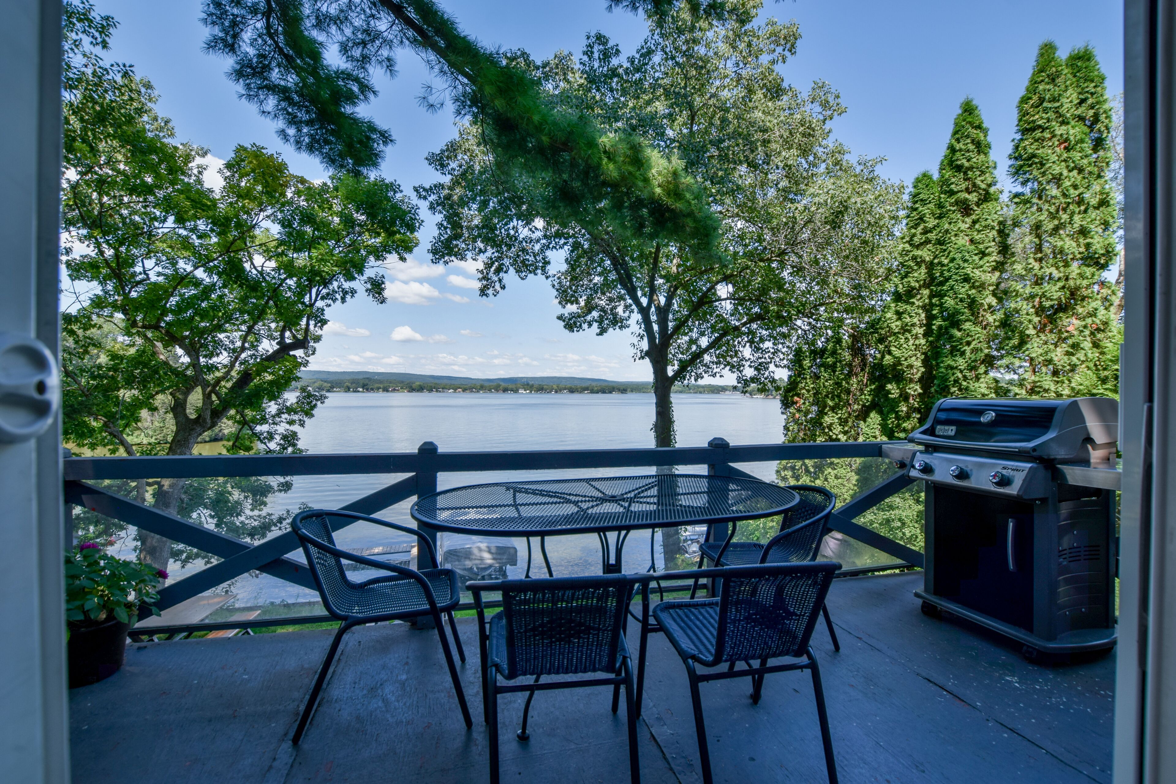 A balcony with a round table and chairs, overlooking a lake with trees and a grill.