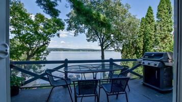 A balcony with a round table and chairs, overlooking a lake with trees and a grill.