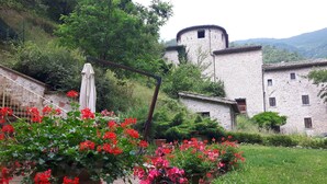 A rustic stone building with a tower, surrounded by lush greenery and flowering plants. - Antica Torre Del Nera (Scheggino)