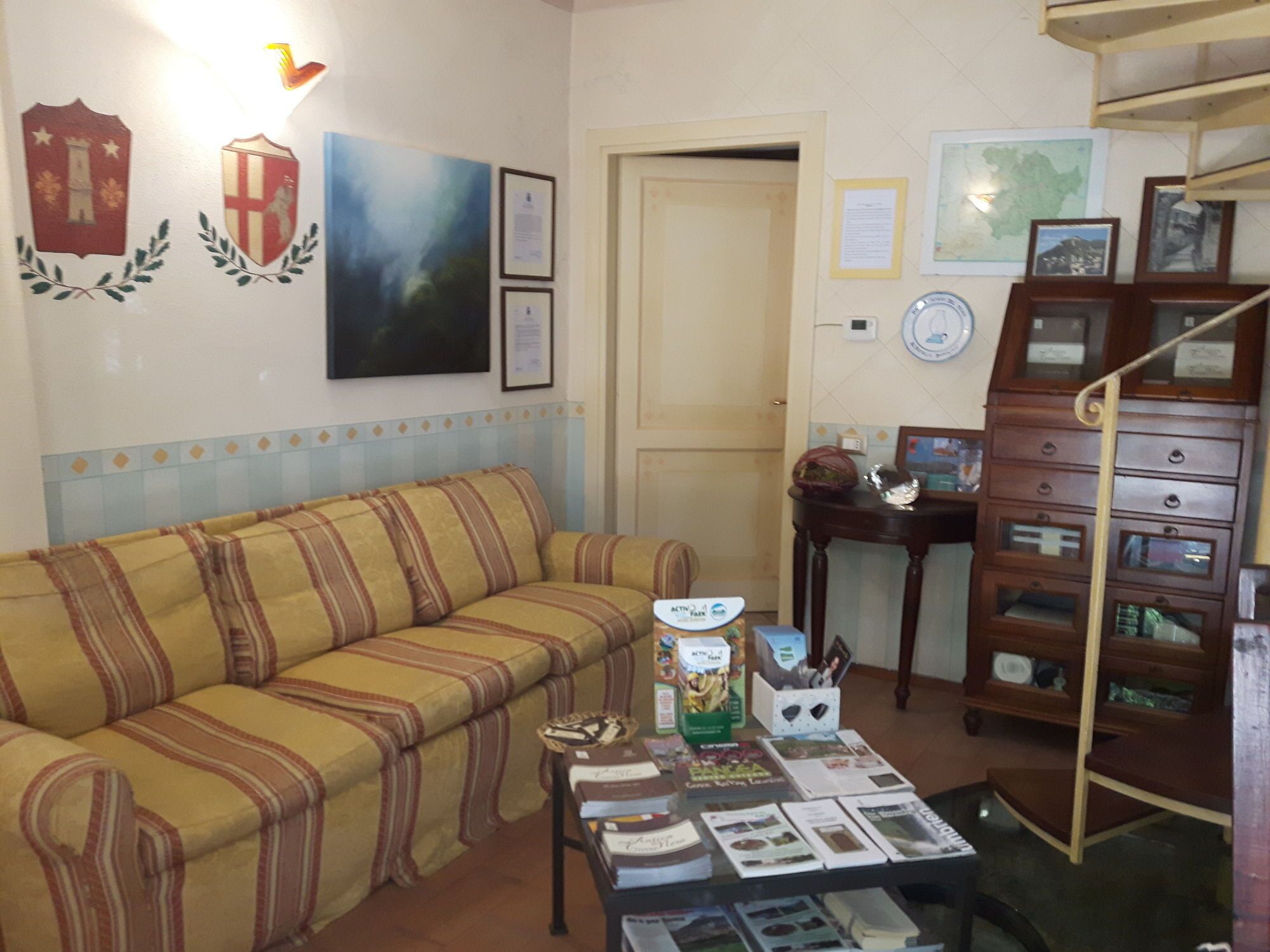 A living room with a striped sofa, a coffee table with magazines, a wooden cabinet with drawers, and wall decorations including a map and framed documents.