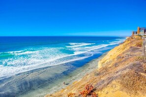 Un paysage côtier offrant une vue dégagée sur l’océan, une plage de sable et une falaise sur laquelle se trouvent des bâtiments.