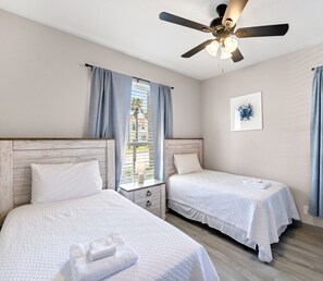 Une chambre d’hôtel avec deux lits simples, un ventilateur de plafond, une fenêtre avec des rideaux, une table de chevet avec une lampe et un tableau encadré au mur.