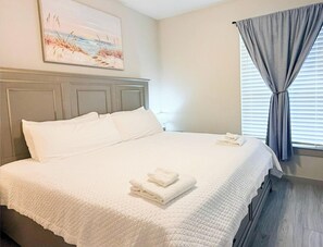 A clean, well-lit bedroom with a neatly made bed, a painting on the wall, and a window with curtains.
