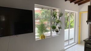 A modern living room with a flat-screen TV mounted on the wall, a large window with potted plants, and a kitchen area with a yellow door.