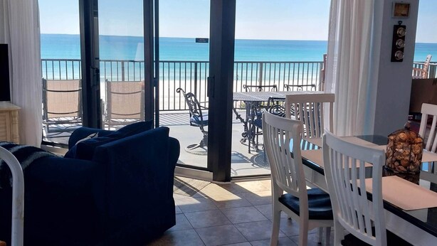 Une pièce dotée d’une grande porte-fenêtre coulissante donnant sur un balcon avec vue sur la plage et l’océan. On y trouve une table à manger blanche avec des chaises, un canapé bleu et un petit téléviseur posé sur un meuble.