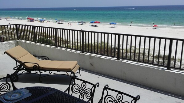A balcony with a lounge chair and table, overlooking a beach with umbrellas and people.