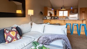 Een moderne woonkamer met een bank, een salontafel, en een keukenhoek met houten kastjes en blauwe krukken.