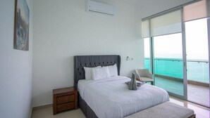 Un dormitorio con una cama grande, una mesita de noche, una silla y vista al océano a través de un ventanal amplio.
