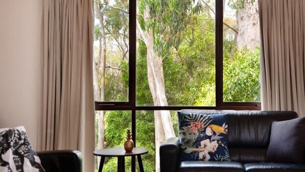 Una sala de estar con un sofá de cuero negro, una mesita redonda con una piña, y vistas a un bosque a través de amplios ventanales.