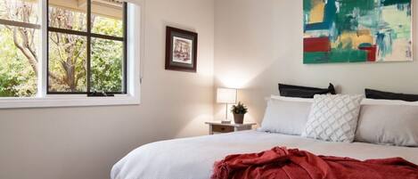 Une chambre à coucher avec un lit, une fenêtre donnant sur des arbres, une table de chevet avec une lampe et un tableau accroché au mur.