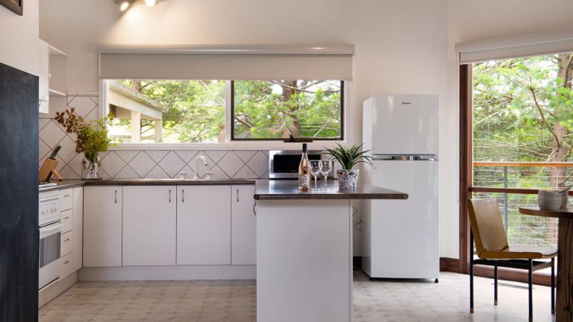 A modern kitchen with white cabinets, a central island, a microwave, and a refrigerator. There is a window with a view of trees and a chair at a small table.