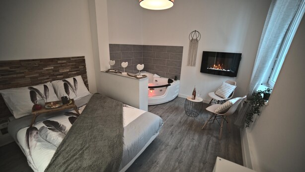 A modern hotel room with a bed, a small seating area with a chair and a table, a fireplace, and a TV mounted on the wall. - LA LOVE LIFE (Mulhouse)