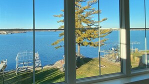 A view through a window with a clear blue sky, a tree, and a body of water with a dock and chairs. - Luxury Lakefront Home w Miles of Lake Views, private pier, lake toys & game room (Pewaukee)