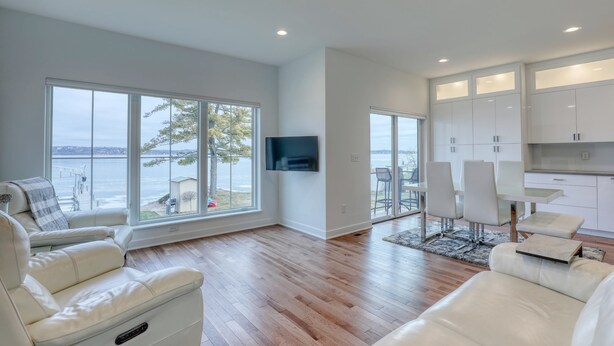 A modern living room with a large window, a flat-screen TV, and a dining area with white furniture. - Luxury Lakefront Home w Miles of Lake Views, private pier, lake toys & game room (Pewaukee)