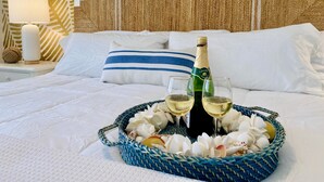 Uma cama com lençóis brancos, um travesseiro azul listrado, uma garrafa de champanhe, dois copos, e uma cesta de vime com conchas e uma fruta amarela.