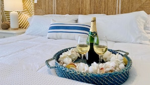 A bed with white linens, a blue-striped pillow, a bottle of champagne, two glasses, and a woven basket with seashells and a yellow fruit. - Sailfish Gulf Suites #4, Garden Views, Steps from the Beach (Holmes Beach)