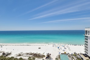 A beach with a clear sky, a white sandy shore, and a building with balconies overlooking the beach.