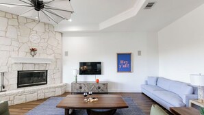 A modern living room with a stone fireplace, a flat-screen TV, a blue wall hanging, and a light blue sofa. - Pool/Spa | 14 Beds | Sleeps 28 - WalkerVR Sundown Escape (Austin)