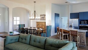 A modern kitchen with blue cabinets, a dining area with striped chairs, and a green sofa in the living room. - Pool/Spa | 14 Beds | Sleeps 28 - WalkerVR Sundown Escape (Austin)