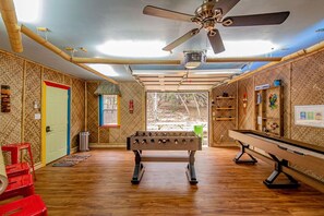 Una habitación con mesa de futbolín, ventilador de techo y ventana con vista a árboles.