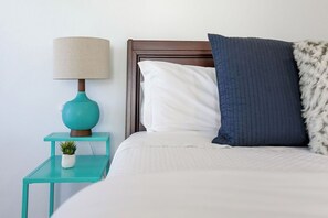 Una cama bien hecha con cabecera de madera, una mesita de noche azul turquesa con lámpara y una pequeña planta en maceta.