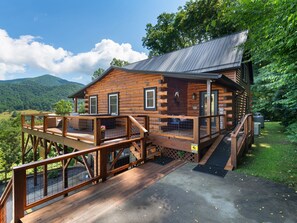 A log cabin with a deck, a covered porch, and a view of the surrounding forest. - Breathtaking Views! Fast WiFi, Pool Table, Hot Tub, lvl 2 EV Charger, Fire Pit! (Maggie Valley)